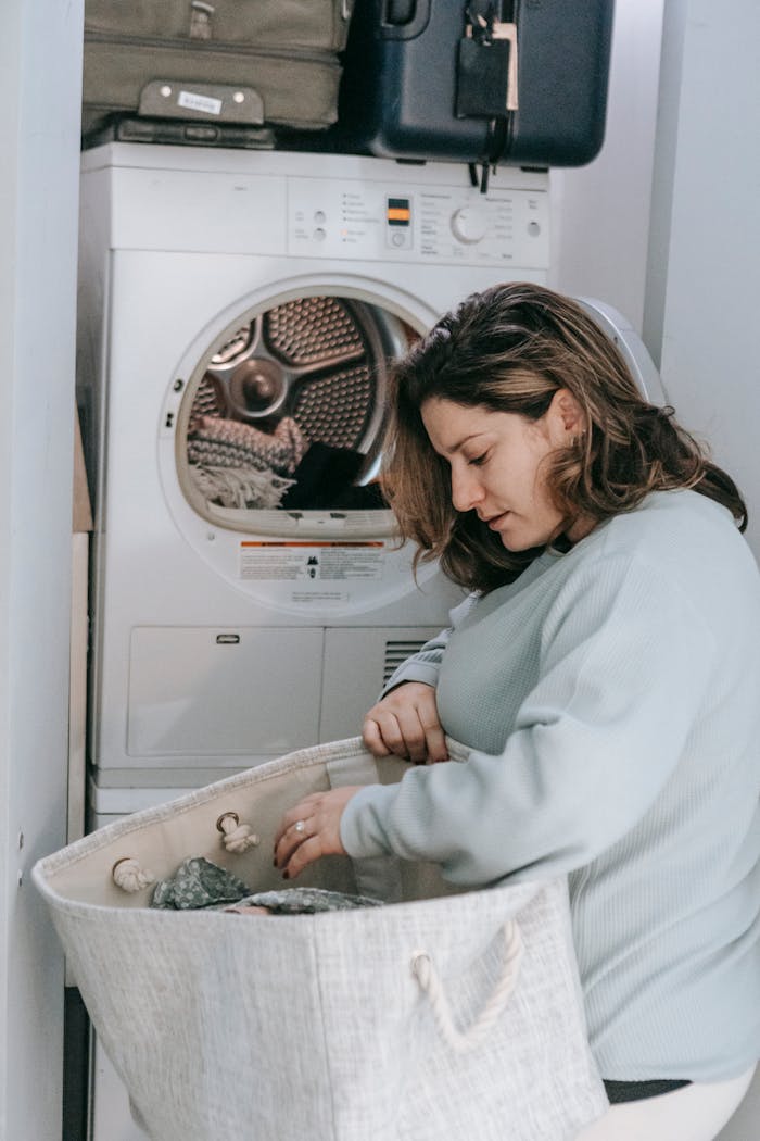 vision Side view of serious female with bag of dirty clothes loading modern washing machine while sitting in light washroom at home